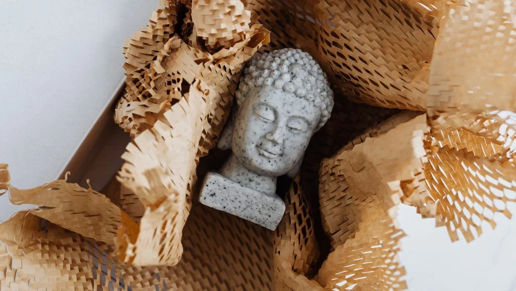 Buddha head sculpture laying in shipping box amongst wrapping paper
